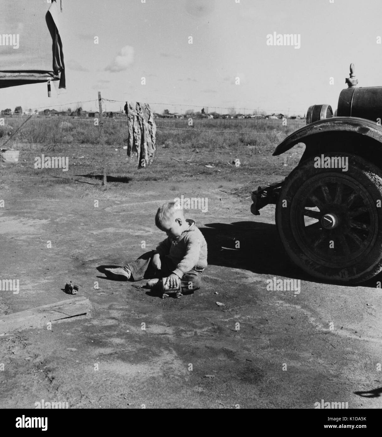 A small boy playing with a handmade toy wagon in the dirt of a migrant camp, near a car, a piece of ruggedly made clothing dries in the background, California, 1936. From the New York Public Library. Stock Photo