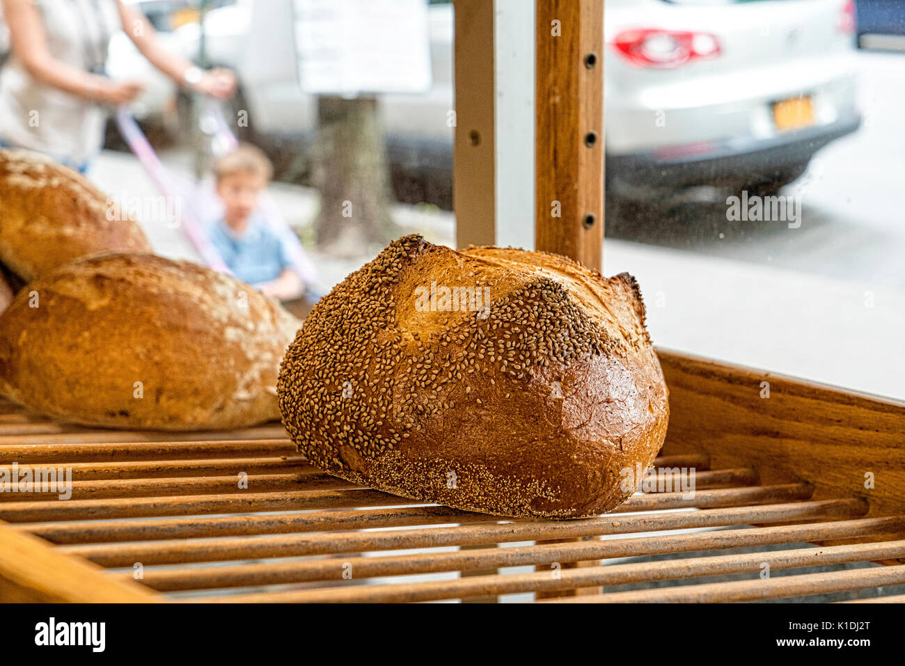 Bronx, NY, USA.  Loaf of Freshly Baked Seeded Semolina Bread, Displayed in a bakersy window on a wooden shelf.  Street, parked cars and woman pushing  Stock Photo