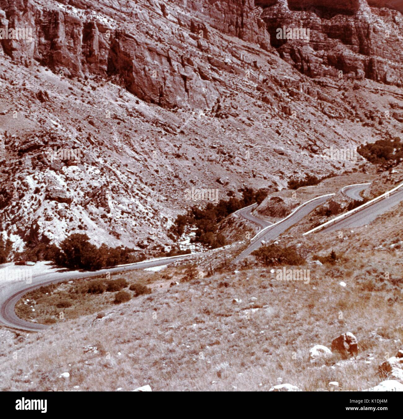 A photograph of several switchbacks along a road leading in to a canyon, taken from the vantage point of a hillside, background consists of a very tall cliff face, Wyoming, 1975. Stock Photo