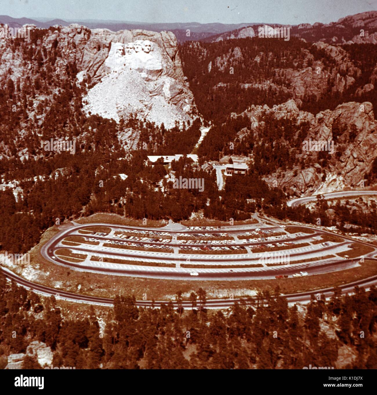 A photograph of Mount Rushmore, taken from a great distance, vantage point was level with the top of the monument, roads leading in and out of the park, along with the parking lot can be seen in the foreground, in the Black Hills in Keystone, South Dakota, 1975. Stock Photo