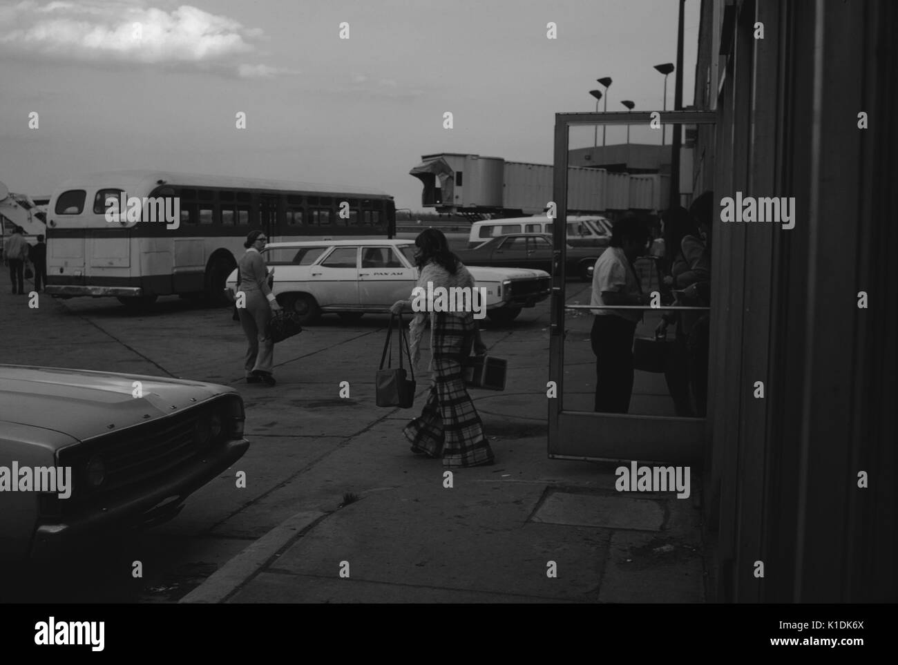 Loading and unloading arrival area in a small airport terminal with two women, one carrying bags and wearing bell bottoms, walking towards a bus, at sunset, 1966. Stock Photo
