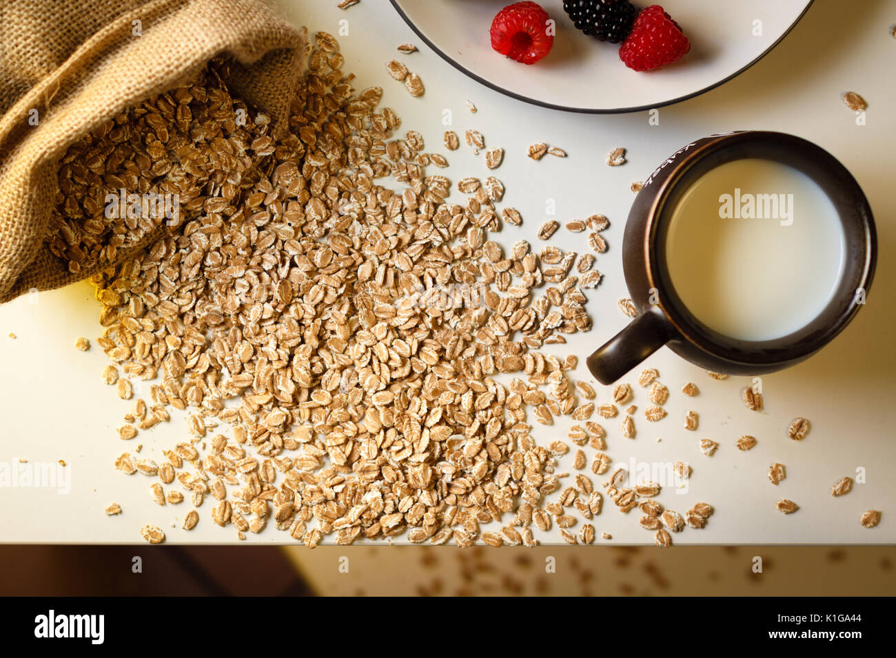 Mess breakfast: scattered oat cereal, milk in cup, blackberry and raspberry on plate, top view. Stock Photo