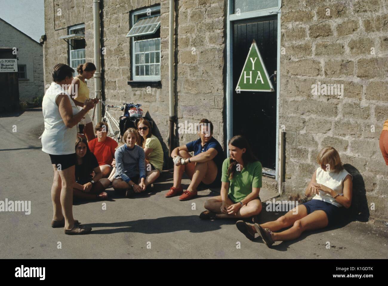 Group of teenage girls sitting outside a youth hostel, a mature woman standing in front of them and speaking to the group, Youth Hostel Association logo visible on the door of the hostel, England, 1966. Stock Photo