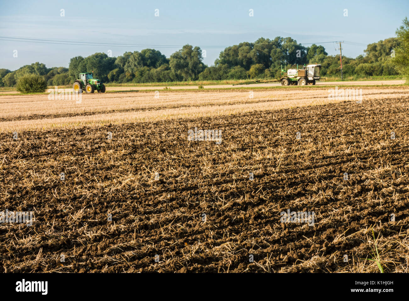A farm field, after a harvested wheat crop, with a tractor turning over the soil at the end of the season, and another vehicle spraying. England, UK. Stock Photo