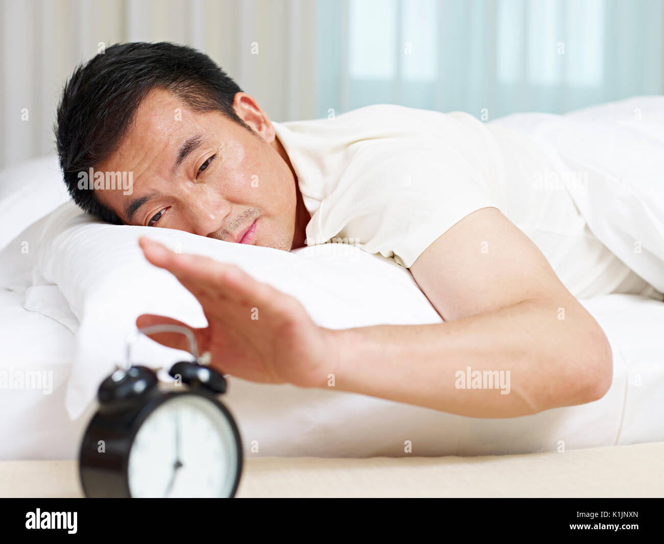 asian man lying in bed trying to stop ringing alarm clock. Stock Photo
