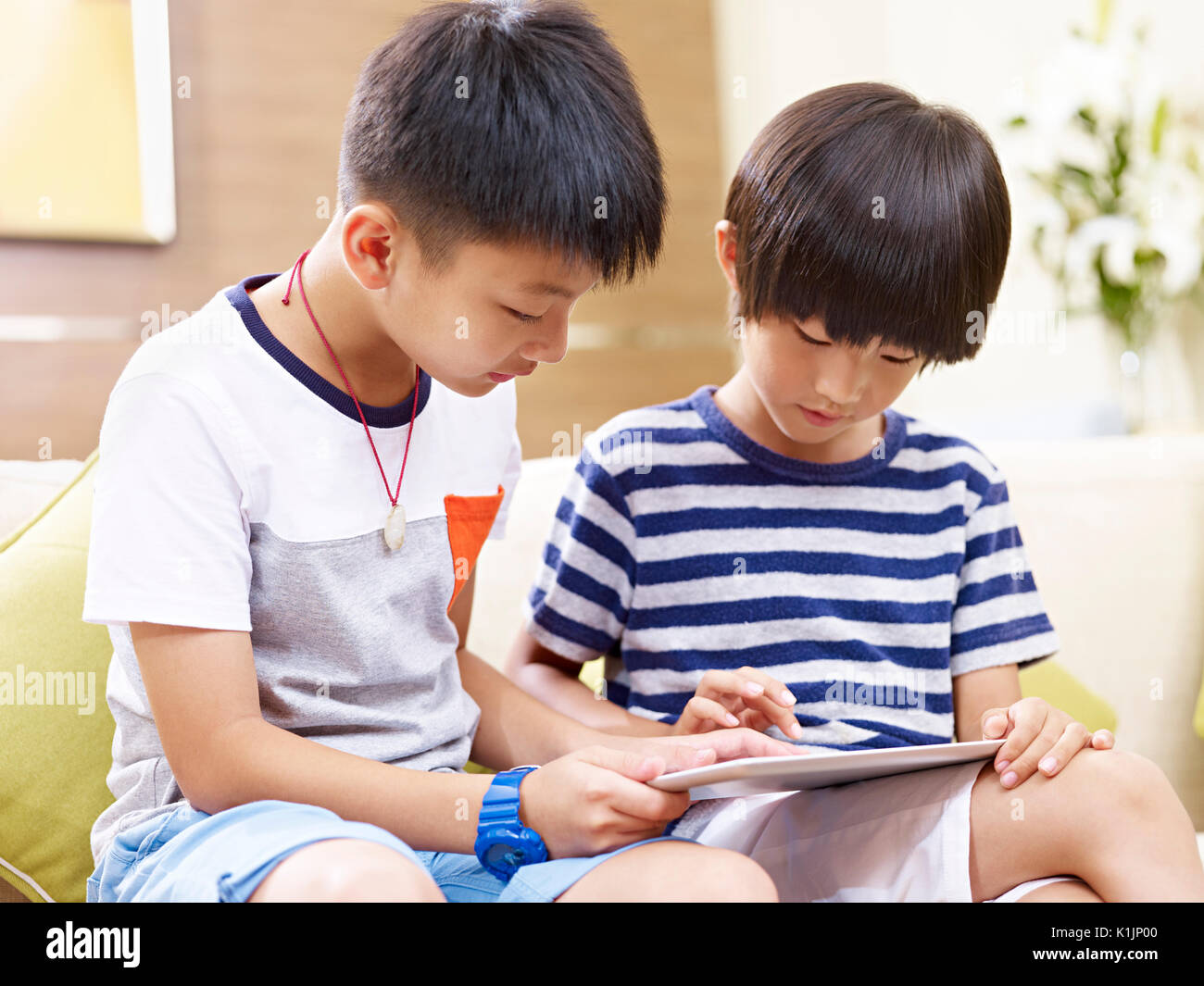 two little asian brothers sitting on couch at home using digital tablet together. Stock Photo