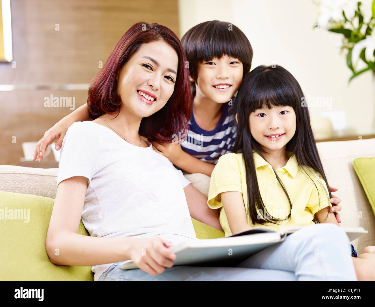 asian mother and two children sitting on couch reading a book. Stock Photo