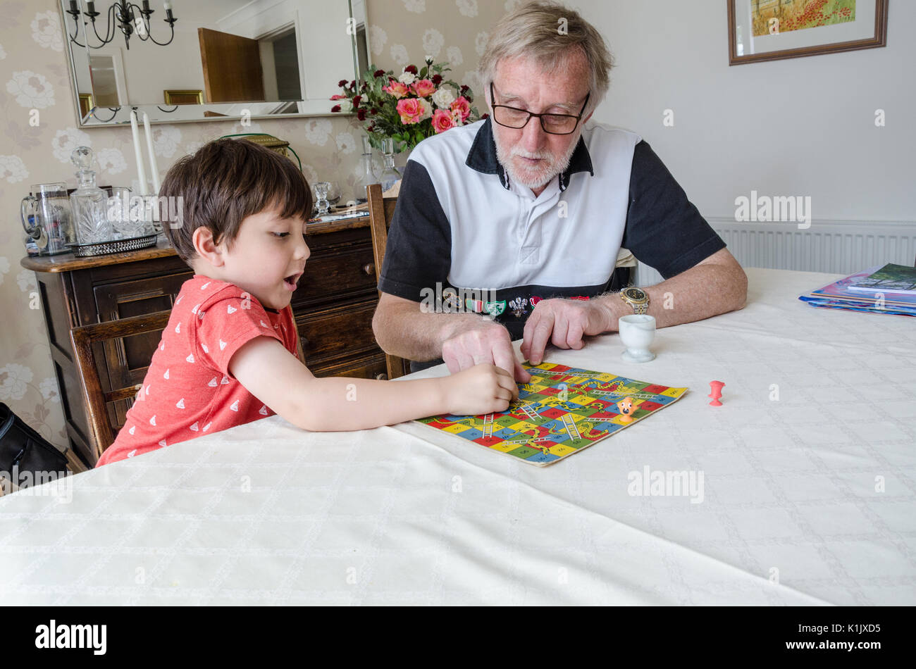 A young boy plays snakes and ladders with his grandfather on the dining room table. Stock Photo