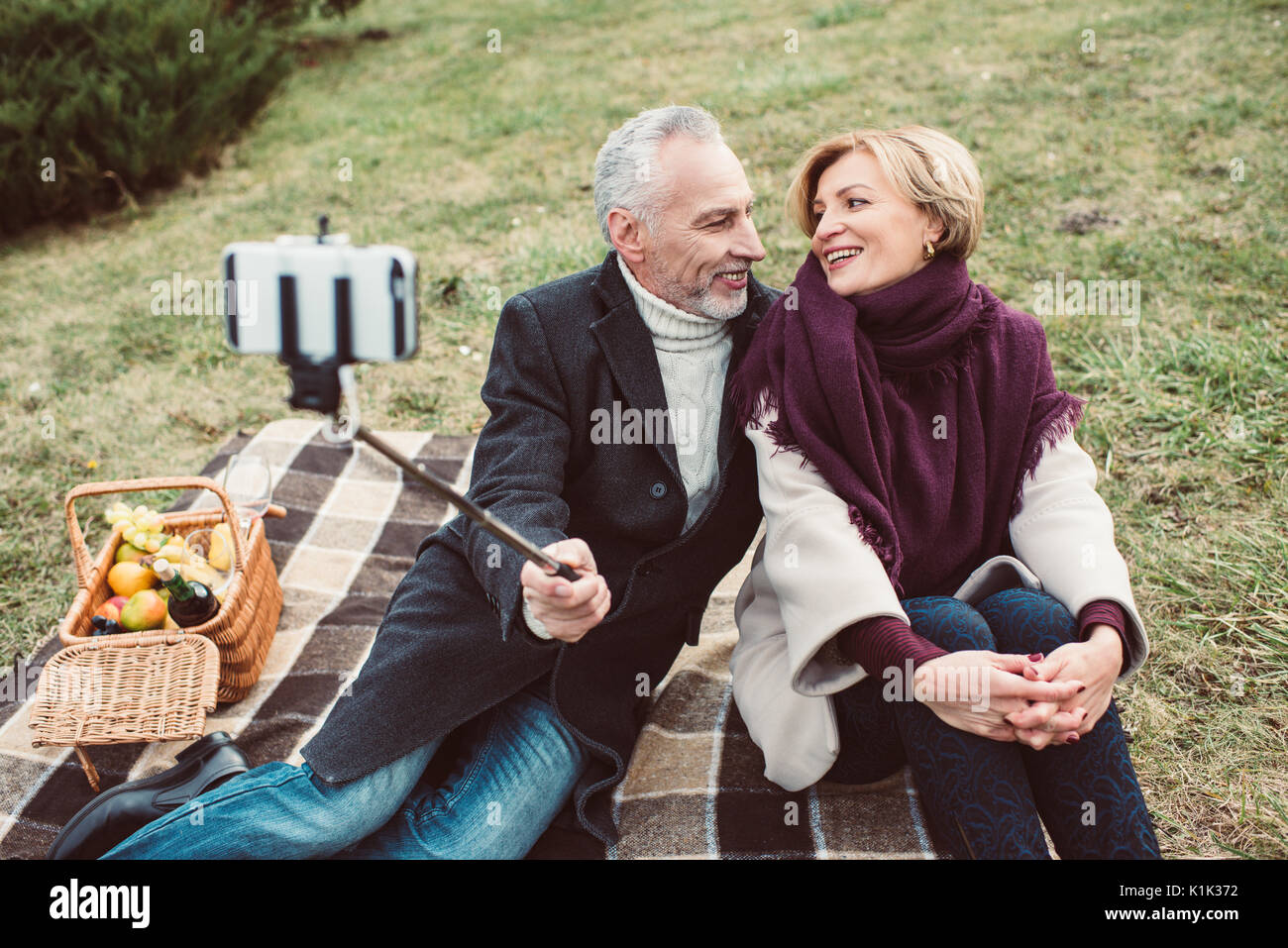 Beautiful smiling mature couple taking selfie and sitting on plaid with picnic basket in autumn park Stock Photo