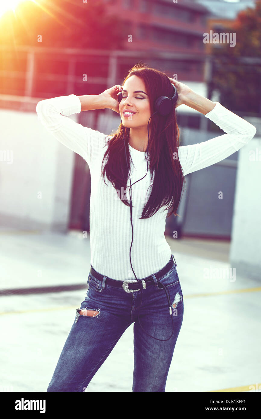 Young trendy woman listening music by headphones, closed eyes, dawn Stock Photo