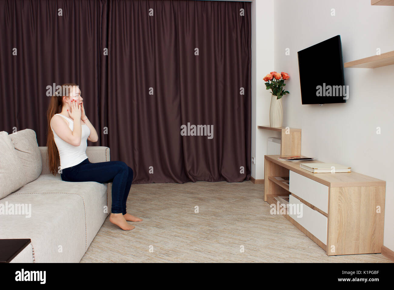 Scared young woman watching tv. Brunette girl sitting on couch With a pillow near the face Stock Photo