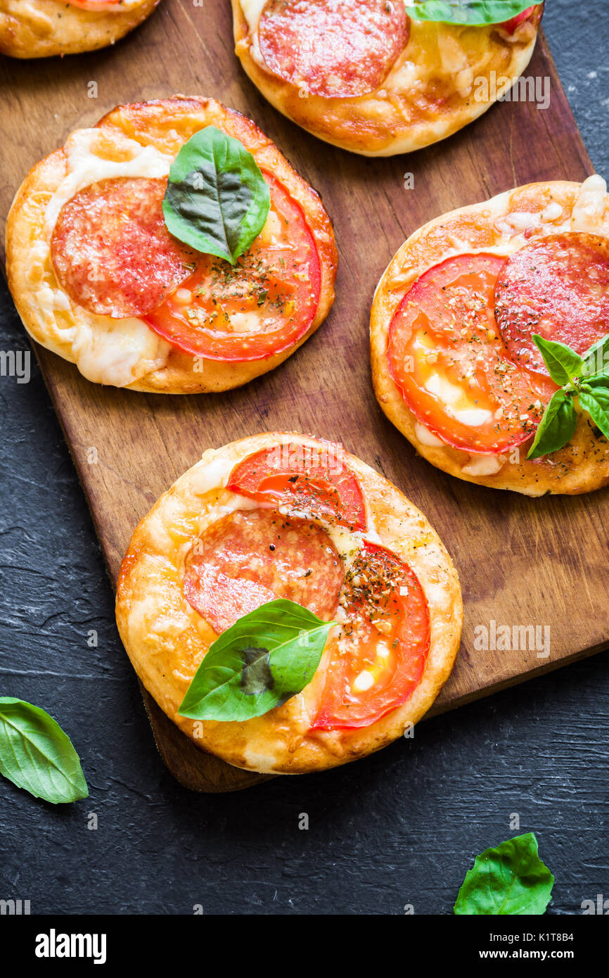 Mini Pizza - Fresh homemade mini pizzas with pepperoni, cheese, tomatoes and basil on rustic black stone background. Stock Photo