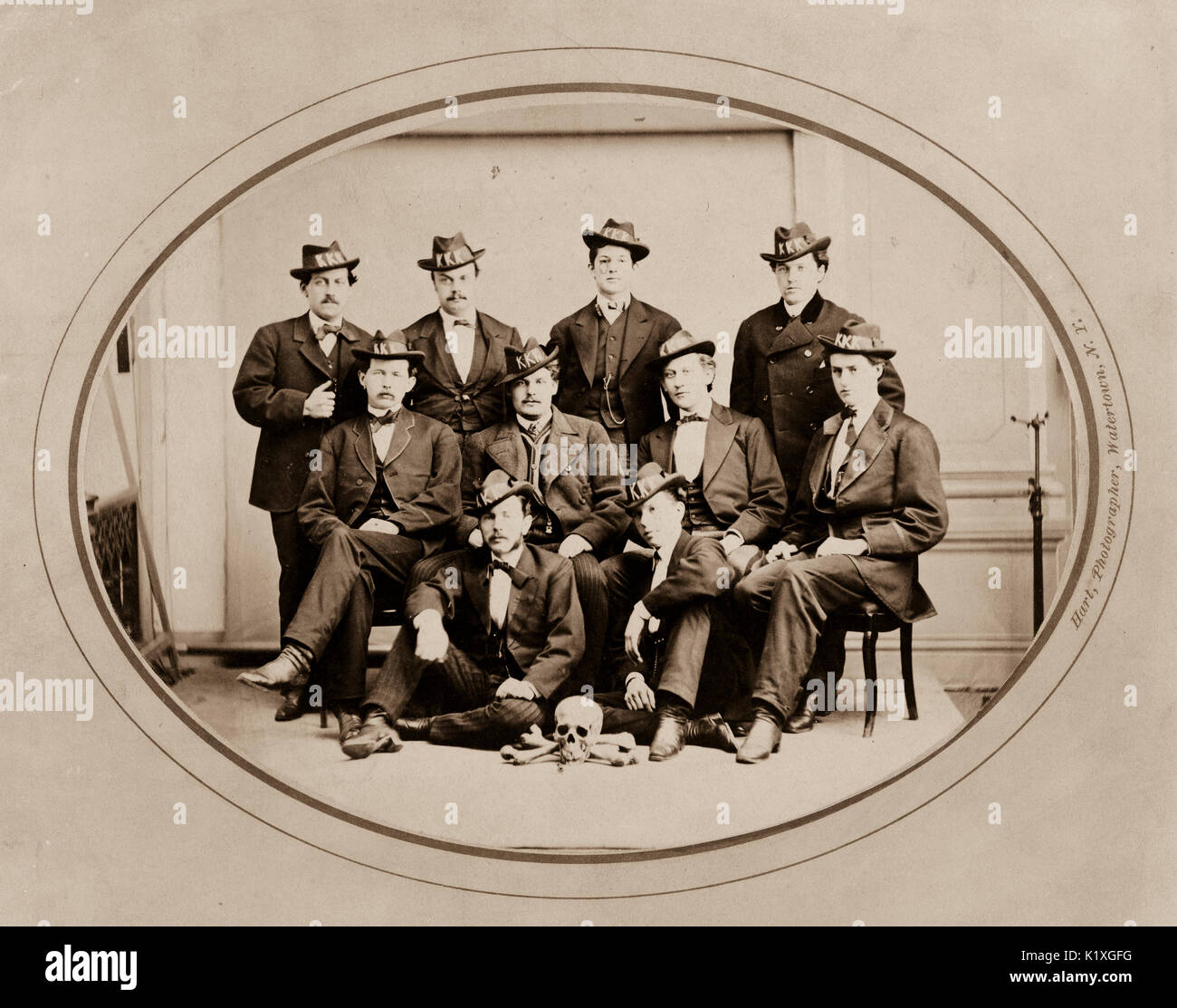 Ku Klux Klan, Watertown Division 289 / Hart, photographer, Watertown, N.Y. Photograph shows ten men posed seated and standing, wearing hats with 'KKK' in large letters, and with a skull and bones arranged on the floor in front of them. Stock Photo
