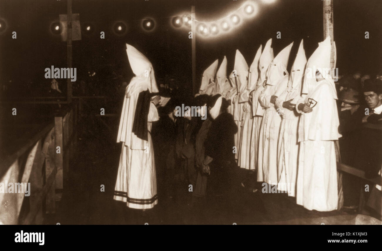 First public appearance of women of the K.K.K. on Long Island, New York. Photograph shows at least four women kneeling in front of shrouded Klansman reading from a book; other Klansmen stand behind them on the platform; spectators watch initiation. Stock Photo