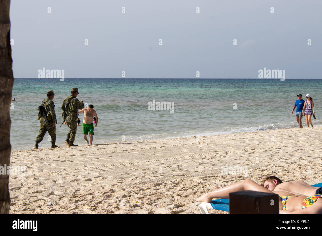 Two armed Mexican soldiers in camouflaged uniforms walking along Caribbean beach with tourists in sim wear ignoring them. Stock Photo