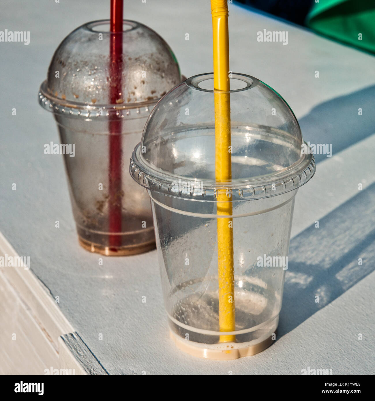Empty plastic glasses with red and yellow straws stand on a white deck. Some green cloth and reflection of blue object in the background. Sunlit abstr Stock Photo