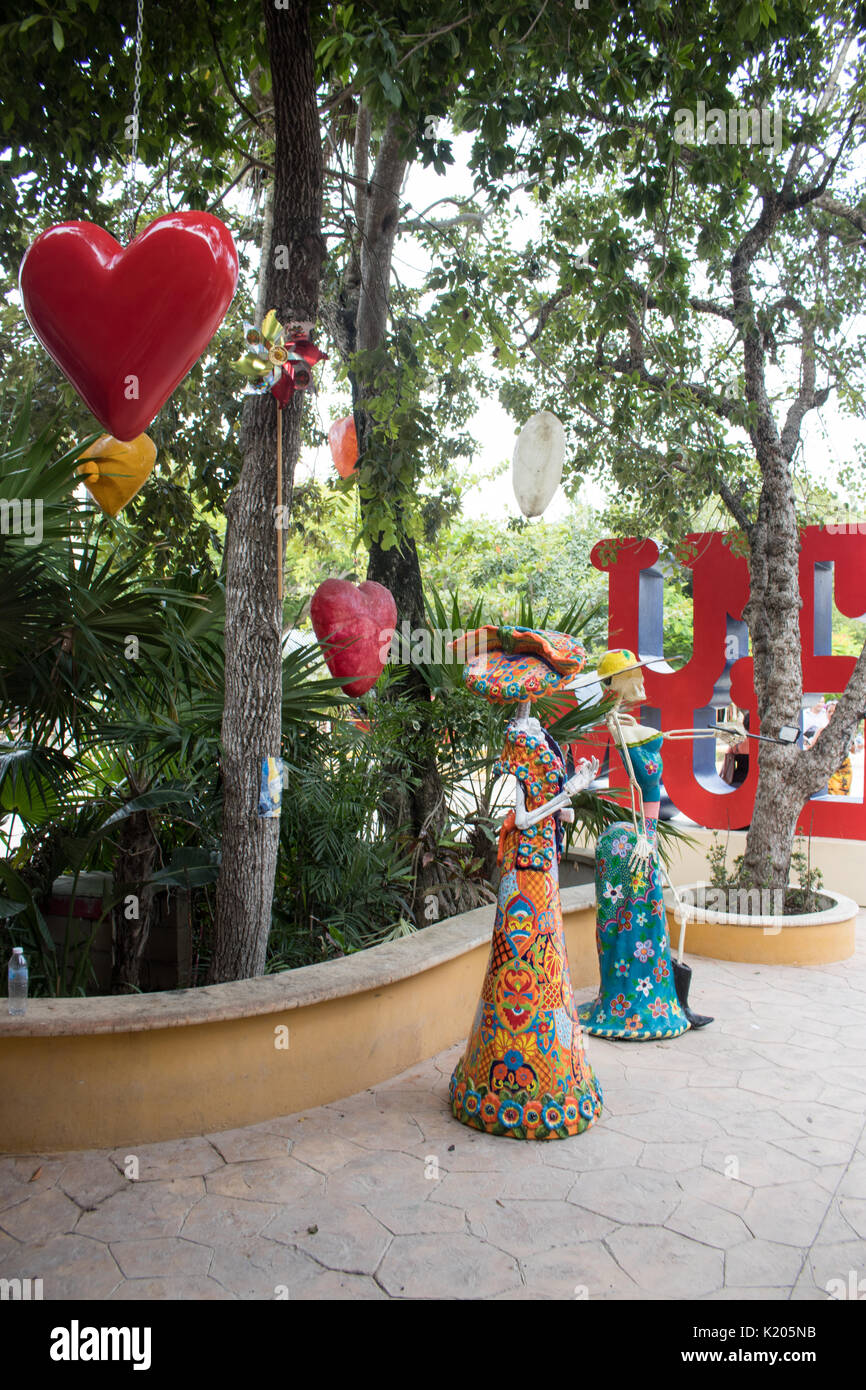 Large red and smaller yellow hearts hanging in trees with Mexican Calaca in bright colored dresses. Stock Photo