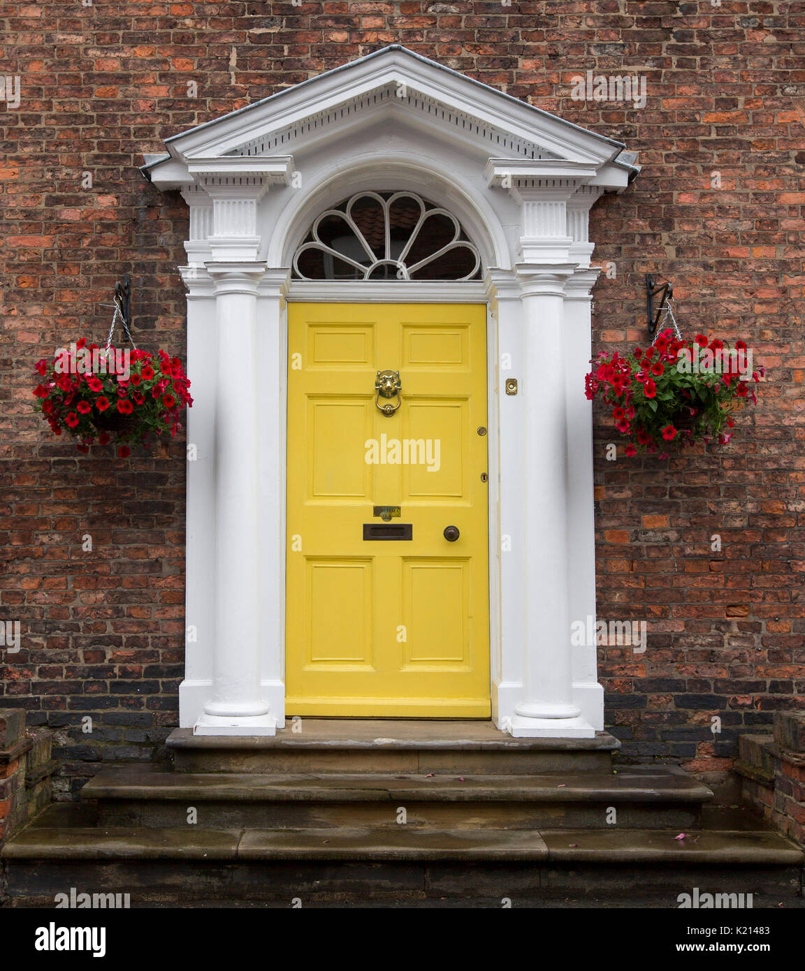 Yellow painted wooden door with white classic style frame / portico bordered by hanging baskets of bright red flowers against dark brick wall Stock Photo