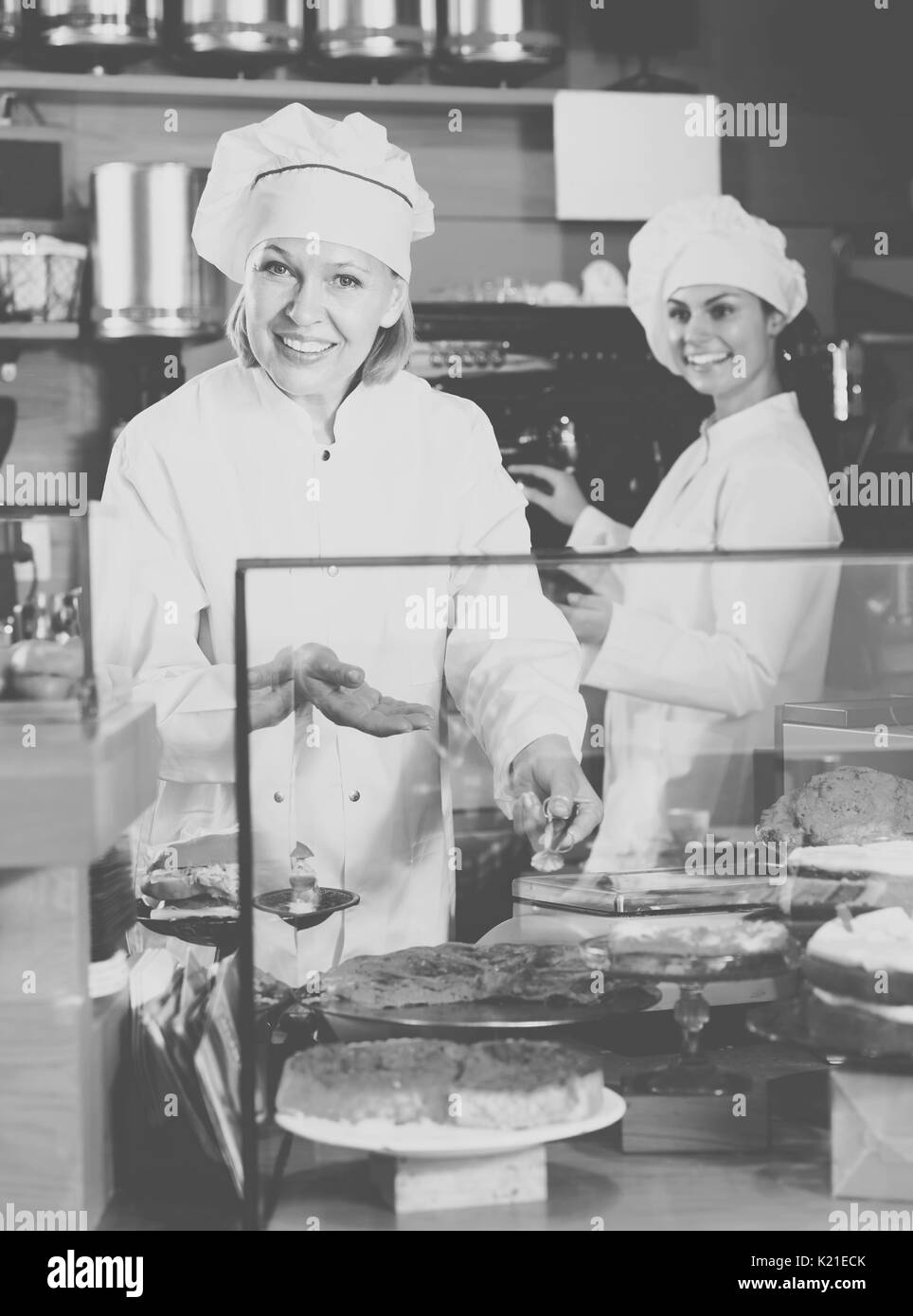 Mature woman and young assistant offering fancy and sponge cakes for sale in cafe Stock Photo