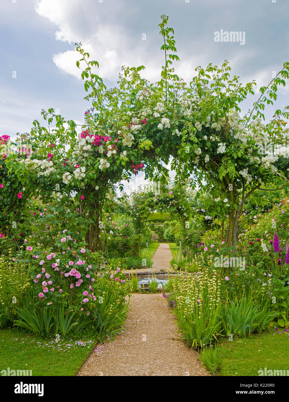 Extensive & stunning English garden with circular pond, herbaceous borders, and pathway leading through archway with climbing roses at Rousham gardens Stock Photo