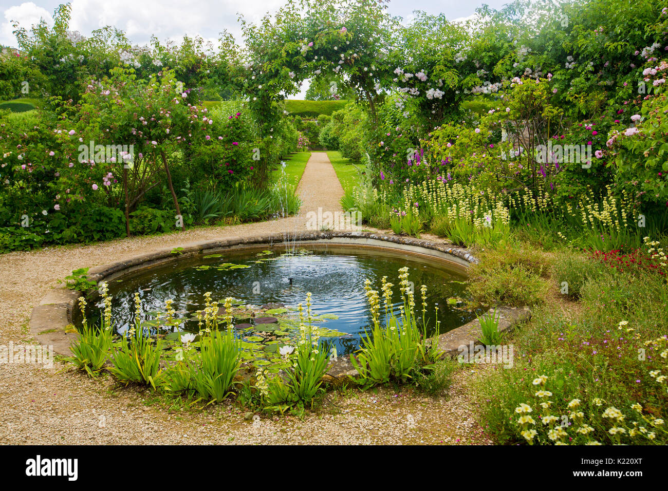Extensive & stunning English garden with circular pond, herbaceous borders, and pathway leading through archway with climbing roses at Rousham gardens Stock Photo
