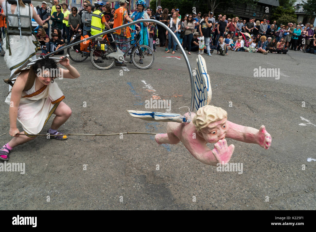 Woman with cherub on a pole that she is having swoop toward to spectators at street's edge. Stock Photo
