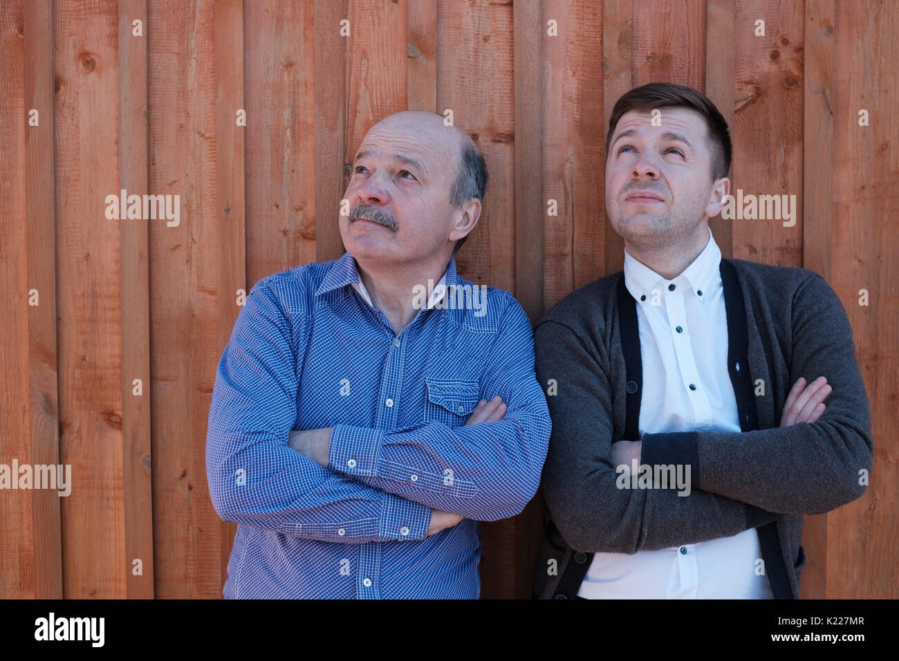 The father and the adult son are standing by the wooden wall and looking thoughtfully upward. Stock Photo