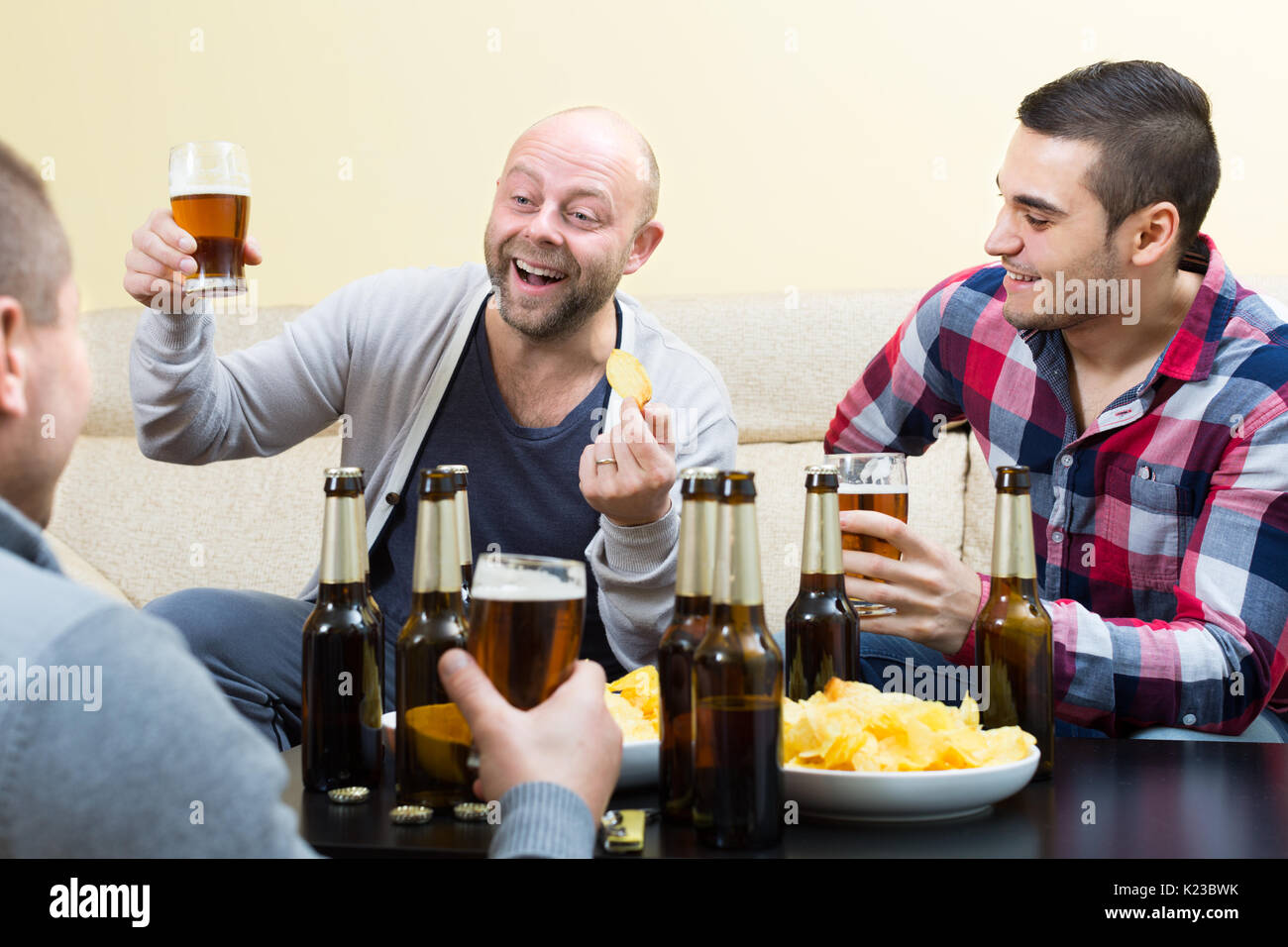 Three happy friends drinking beer  with crisps and talking at home Stock Photo