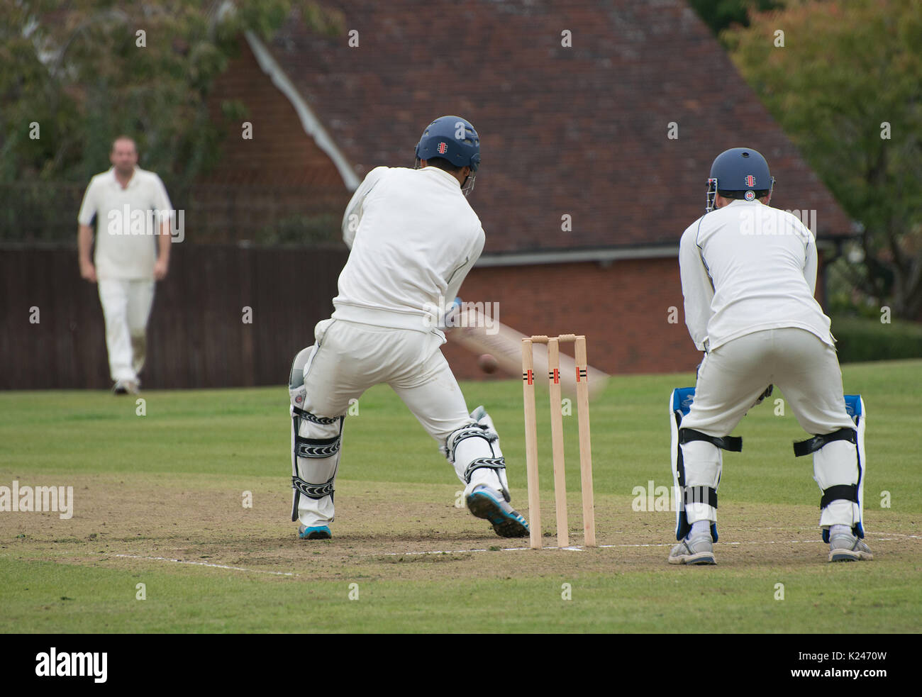 Village cricket match viewed from behind stumps with batsman playing a stroke and wicket keeper ready and with the ball clearly seen Stock Photo