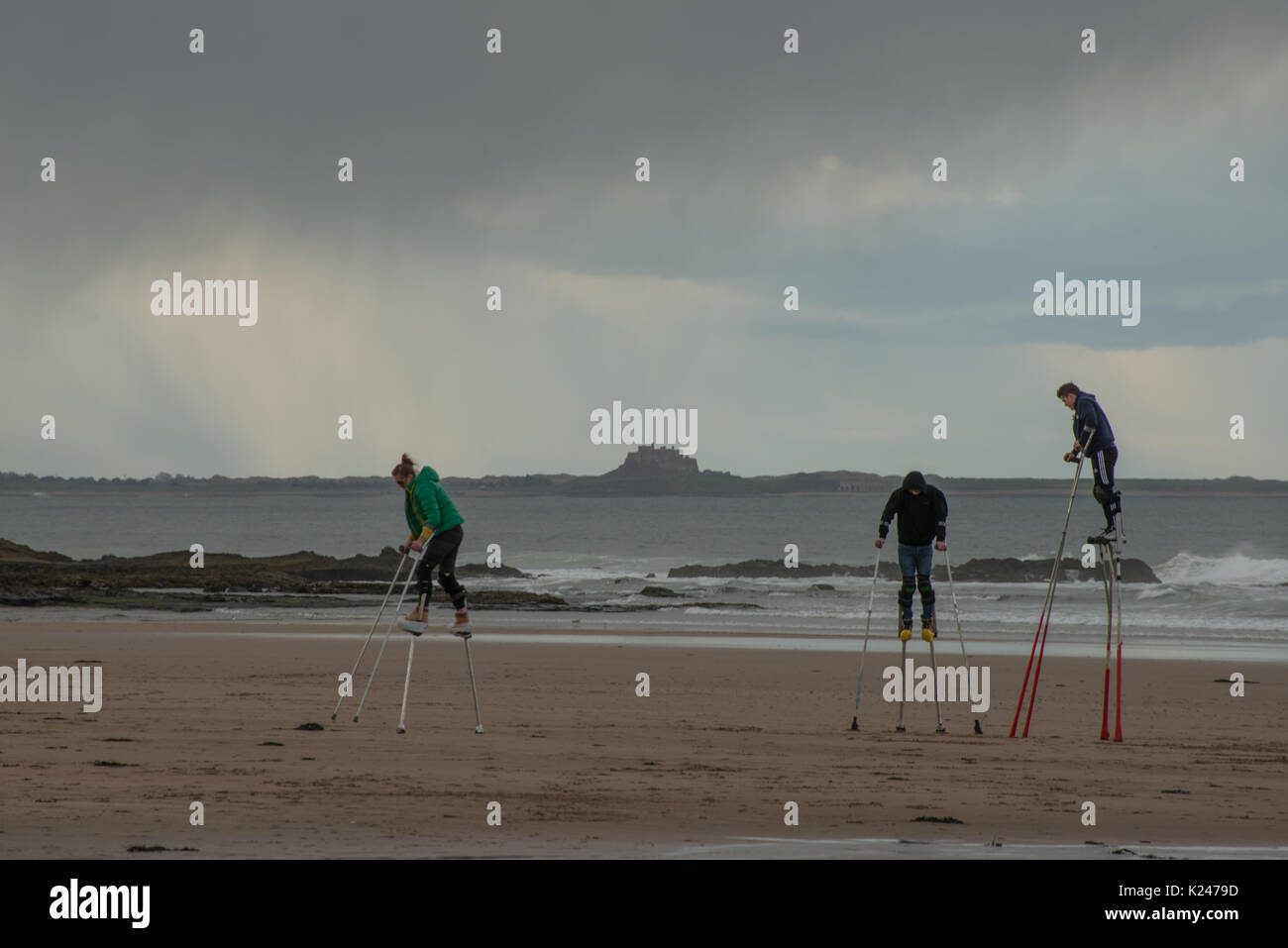 Three stilt walkers in practice on the beach in Northumberland as rain approaches and  with Bamburgh castle in background Stock Photo