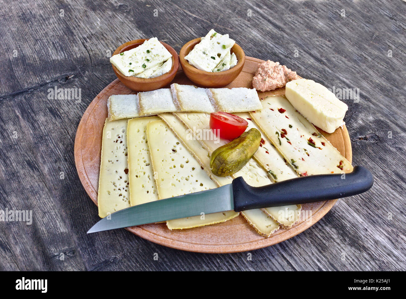 Wooden plate with a selection of cheeses, tomato, pickle and knife on a weathered wooden table Stock Photo