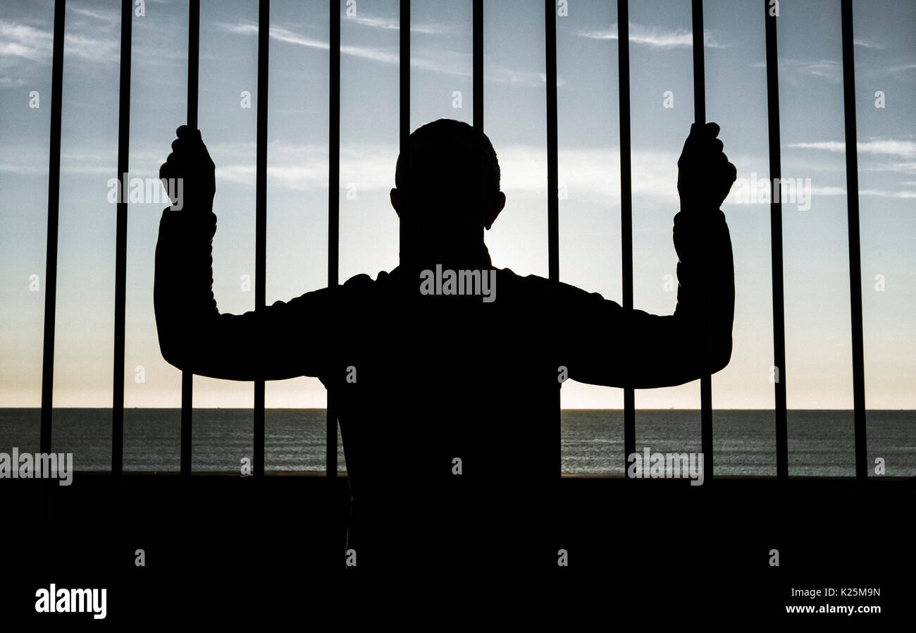 Silhouette of man looking through steel bars with sea in background. Depression, mental health, asylum seeker, immigration... concept image. Stock Photo