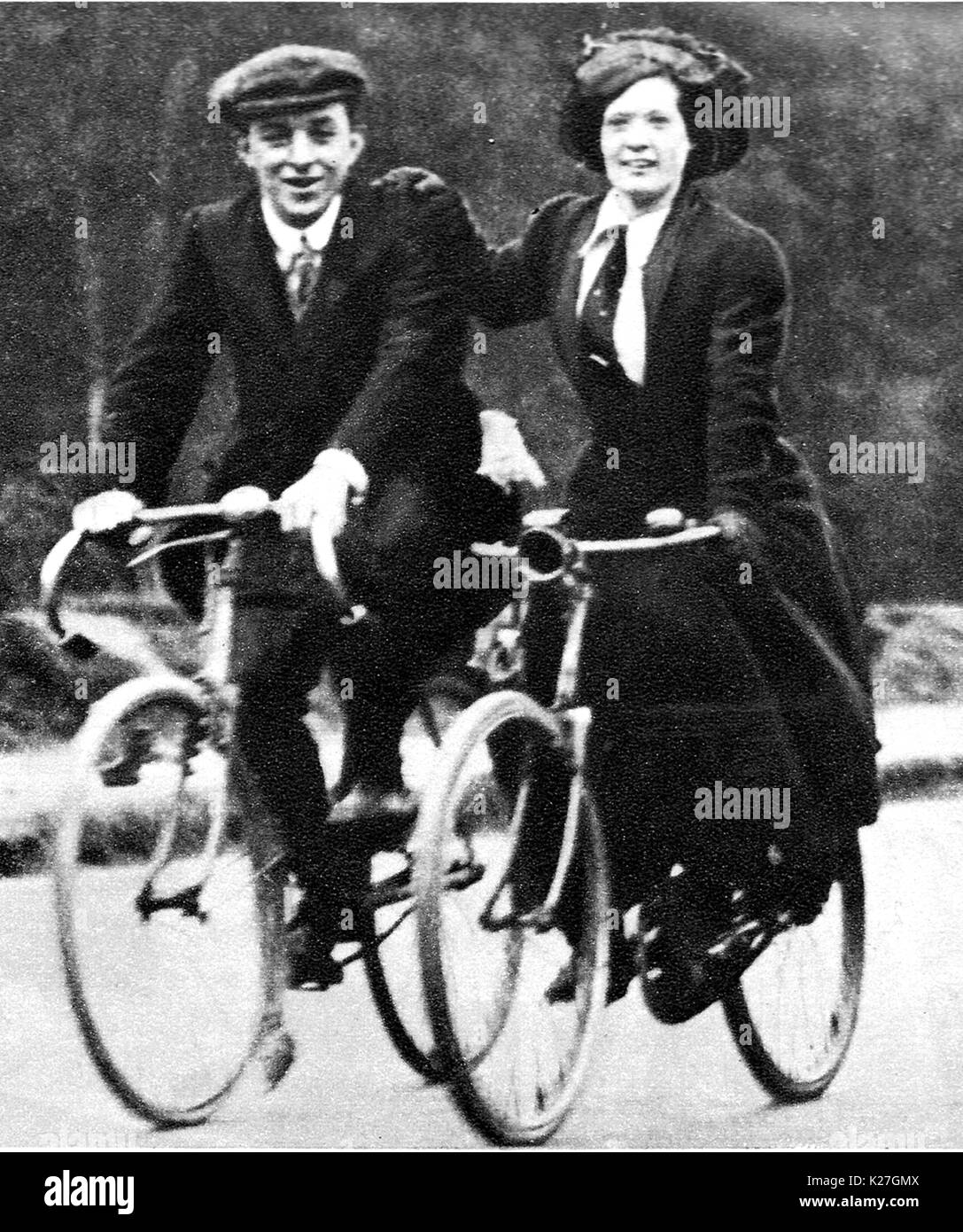 A couple cycling on male & female cycles and wearing the clothing of the time including a flat hat for him and ties for them both Stock Photo