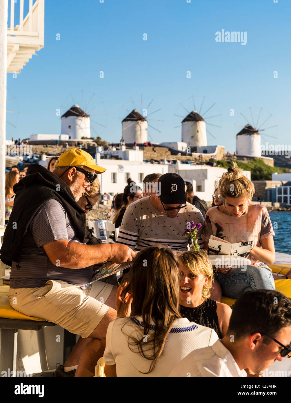 Tourists sitting at a waterfront cafe with the Kato Mili windmills behind them, Mykonos, Greece. Stock Photo