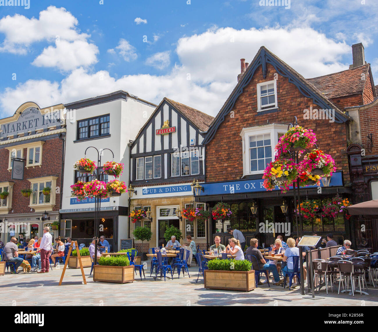 Diners at outdoor tables beside Ox Row Inn in historic market square adorned with hanging baskets of flowers under blue sky in Salisbury, England Stock Photo