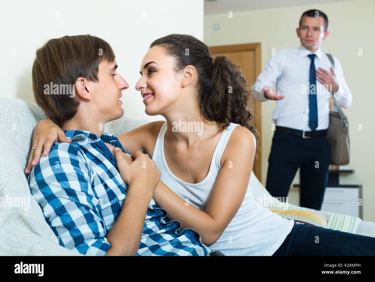 Jealous young husband catching his attractive wife with lover on couch Stock Photo image