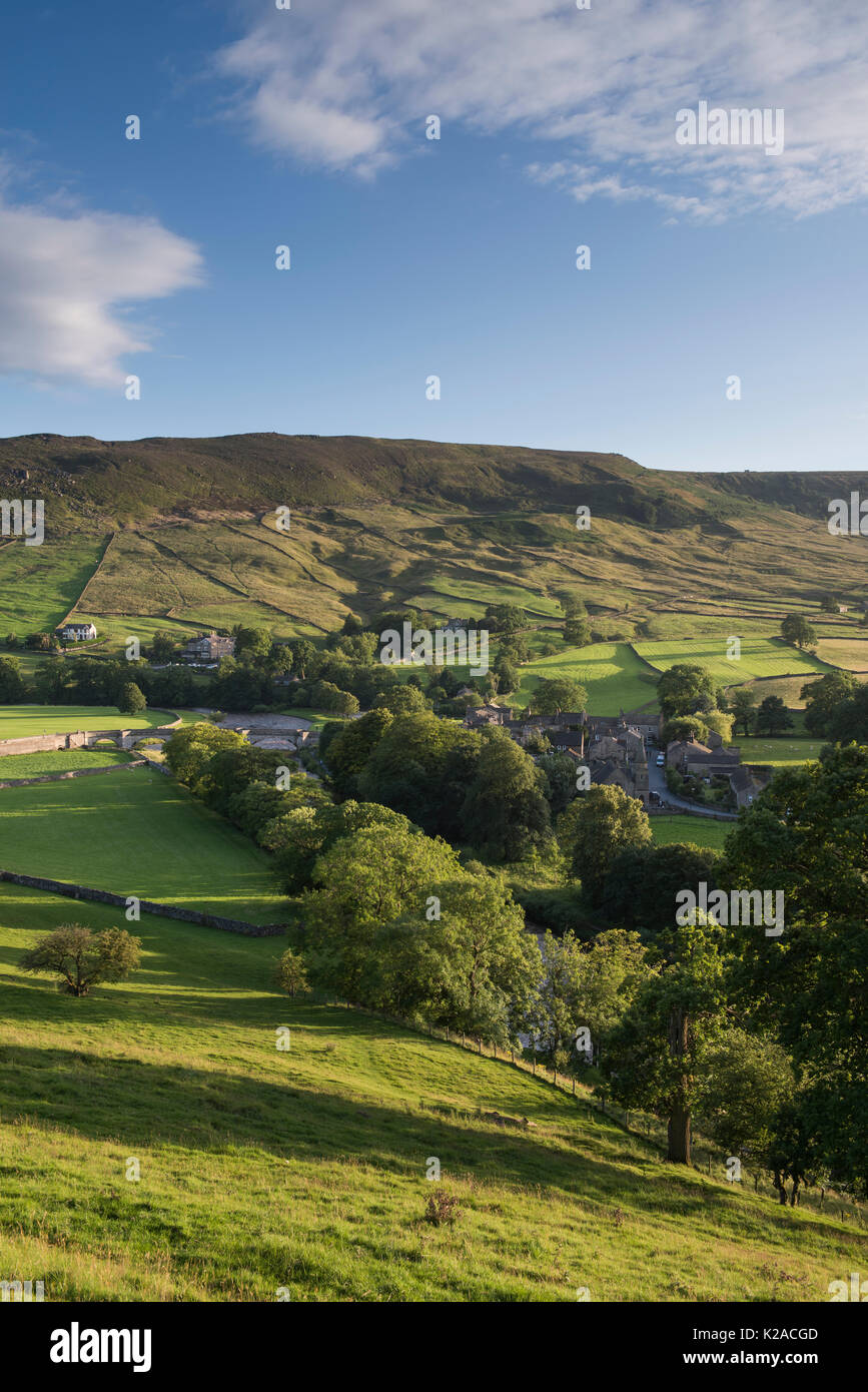 High view over beautiful, sunny Yorkshire Dales village of Burnsall in valley by River Wharfe & with steep, green, hillside beyond - England, GB, UK. Stock Photo