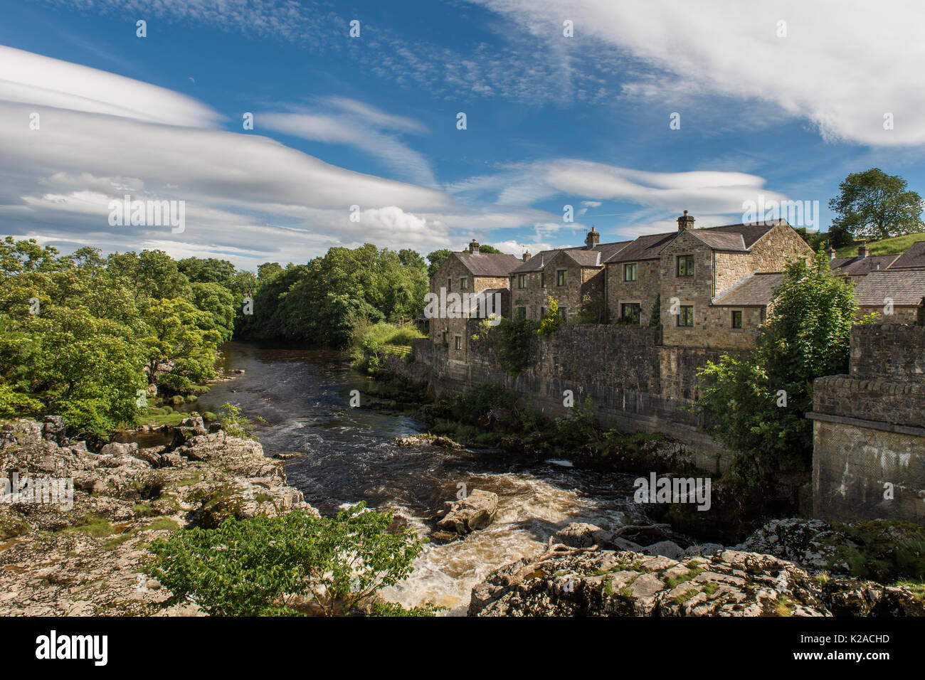 Blue sky over cascading water & riverside houses at sunny, scenic Linton Falls waterfall over River Wharfe, Grassington, Yorkshire Dales, England, UK. Stock Photo