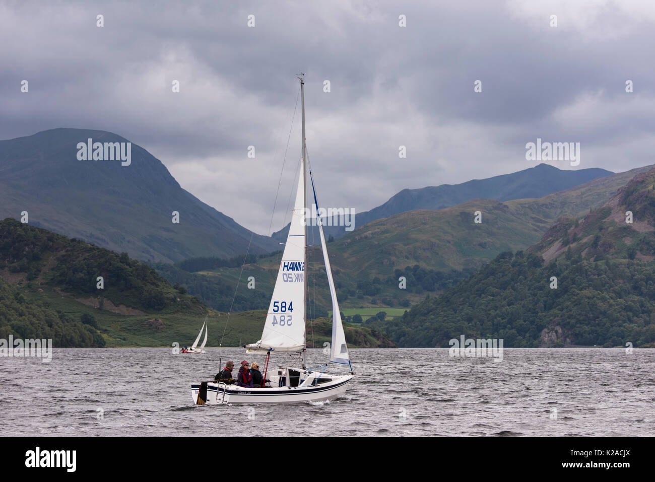 By rolling hills & under grey sky, people are sailing in small  boats on Ullswater - view to Glenridding & Patterdale, Lake District, England, UK. Stock Photo