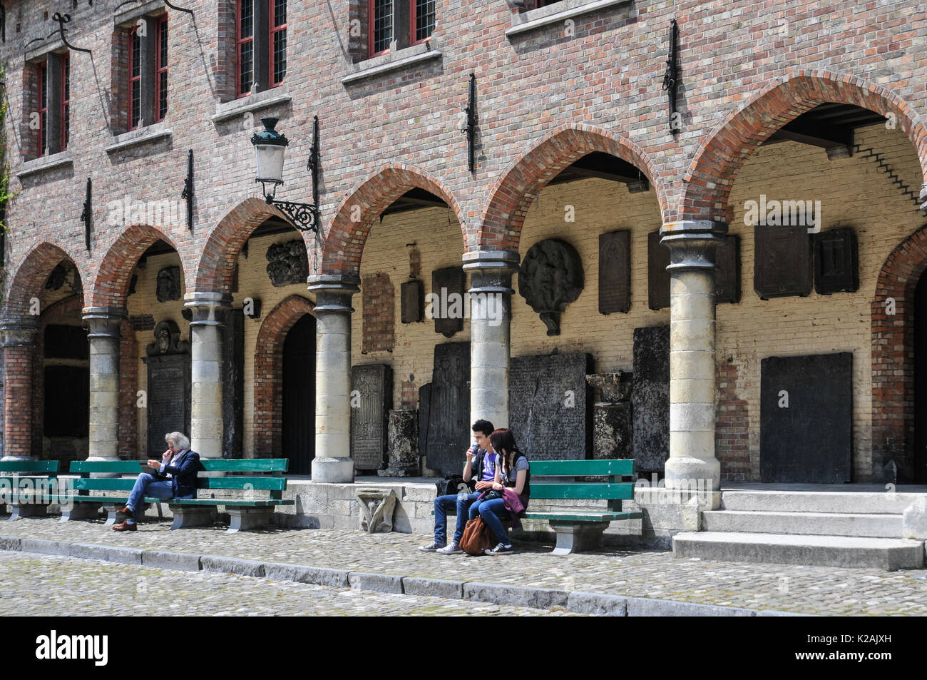 Three people enjoying sitting in the sunshine on benches outside the medieval arches of the Gruuthusemuseum in the city of Brugge / Bruges in Belgium Stock Photo