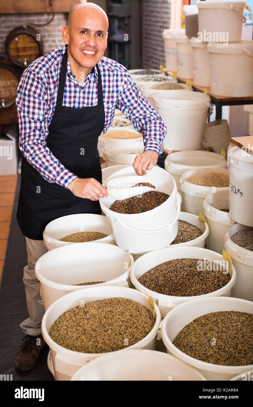 mature glad male seller taking bird seeds with scoop from bucket on market stall Stock Photo