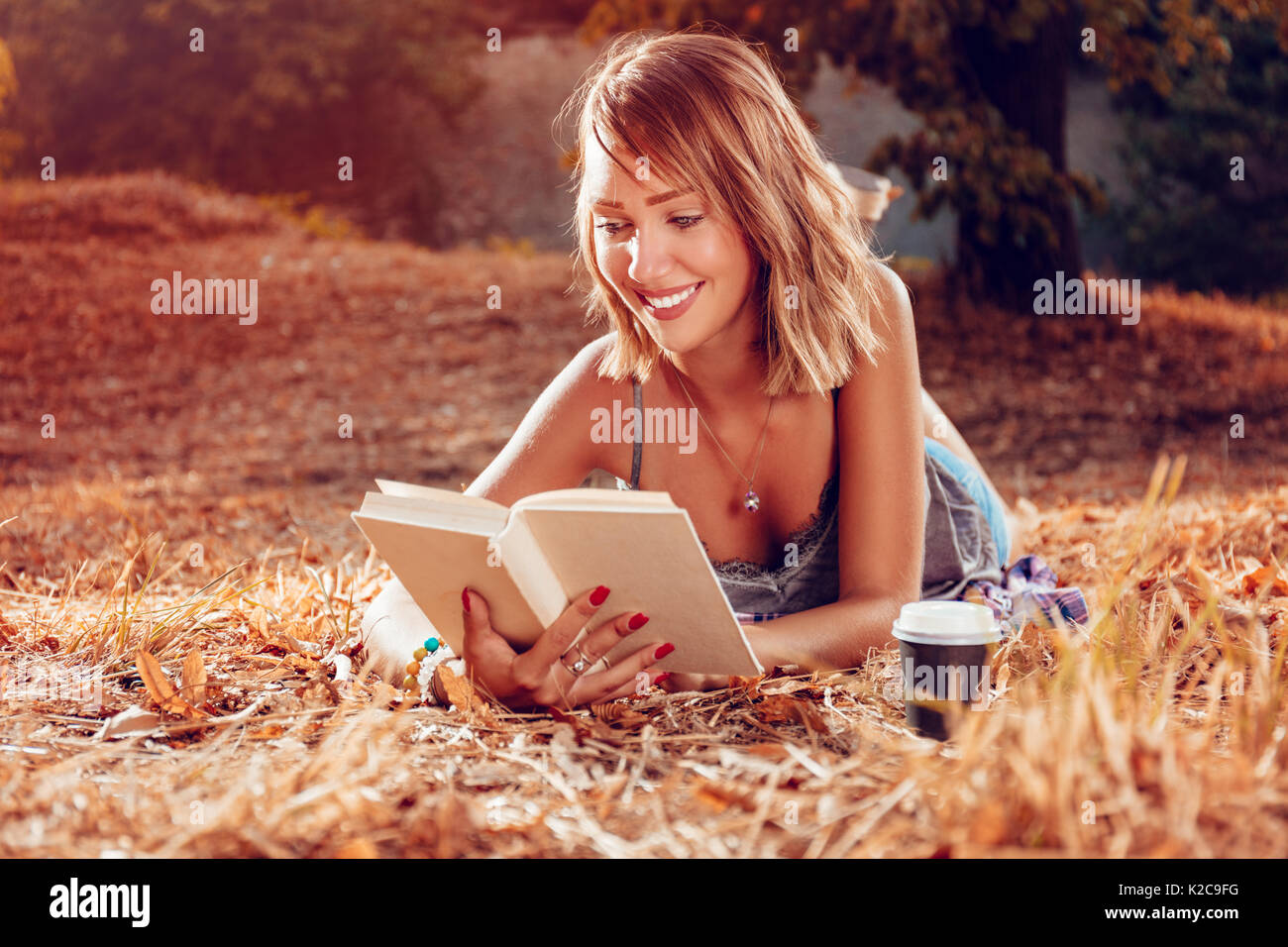 Cute smiling young woman lying on the withered grasses in autumn and reading book. Stock Photo