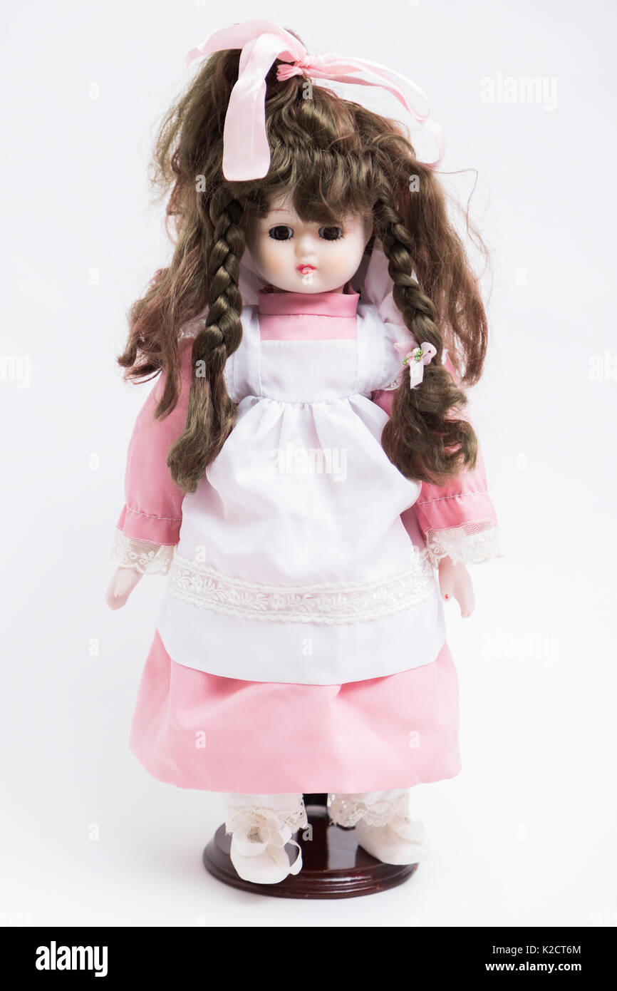 Portrait of ceramic porcelain handmade vintage brunette doll with two pigtails in old pink textile silk dress and white apron with embroidery, bow on  Stock Photo