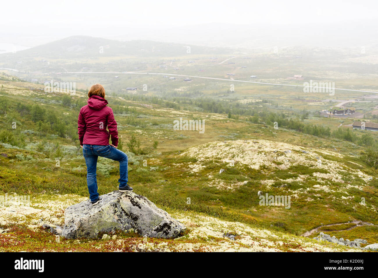 Young adult person looking out over foggy mountain view with small village and road. Location Vasstulan, Norway. Stock Photo