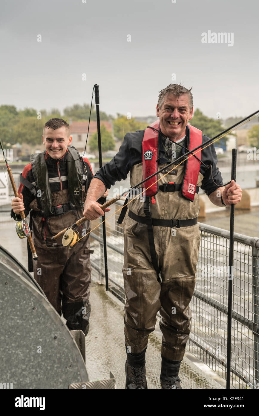 Galway, Ireland - August 5, 2017: Closeup of two laughing fishermen with gear and waders fleeing heavy rain over Corrib River control dam. Gray sky an Stock Photo