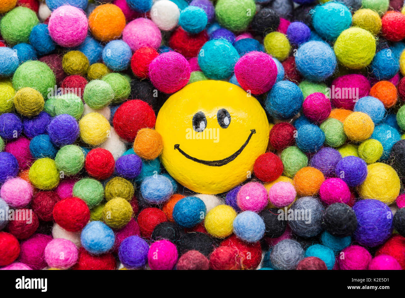 Smiley face made of stone, painted yellow, laughing, lies in between colorful felt balls Stock Photo