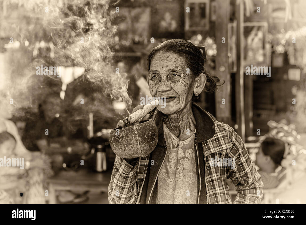 Happy and old wrinkled woman smokes  a big cheroot cigar in public. Vintage black and white processed. Stock Photo