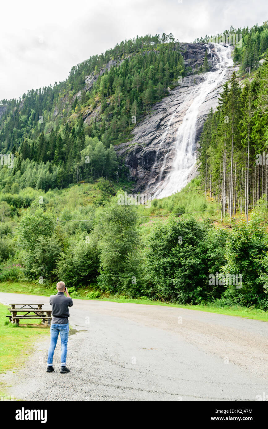 Young adult man standing below woodland waterfall taking pictures with mobile phone. Water cascading over the mountainside. Location Reiarsfossen in s Stock Photo