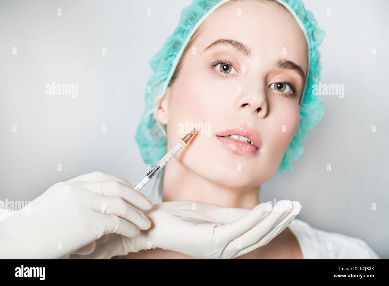 Doctor aesthetician makes hyaluronic acid beauty injections in the nasolabial fold of female patient in a green medical cap Stock Photo