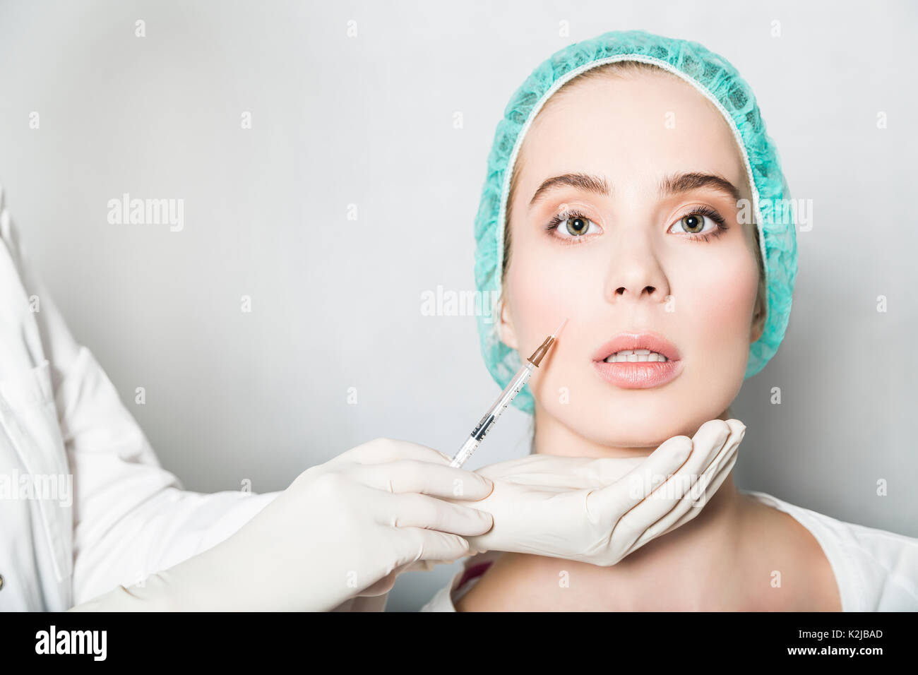 Doctor aesthetician makes hyaluronic acid beauty injections in the nasolabial fold of young female patient in a green medical cap Stock Photo